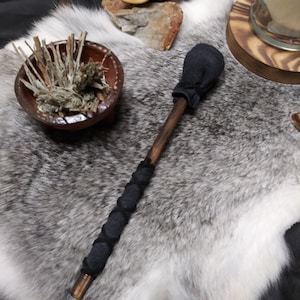 May include: A handcrafted wooden smudge stick holder with a dark handle and a black fabric head. The handle is wrapped with black fabric. A small ceramic bowl with dried herbs is also visible.
