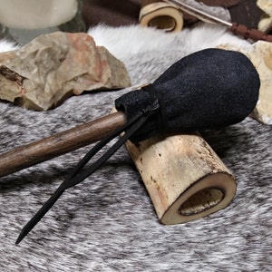 May include: A handcrafted wooden club with a black suede pouch attached. The club features a dark brown handle and is adorned with black leather accents. The background includes rocks and a fur-like surface.