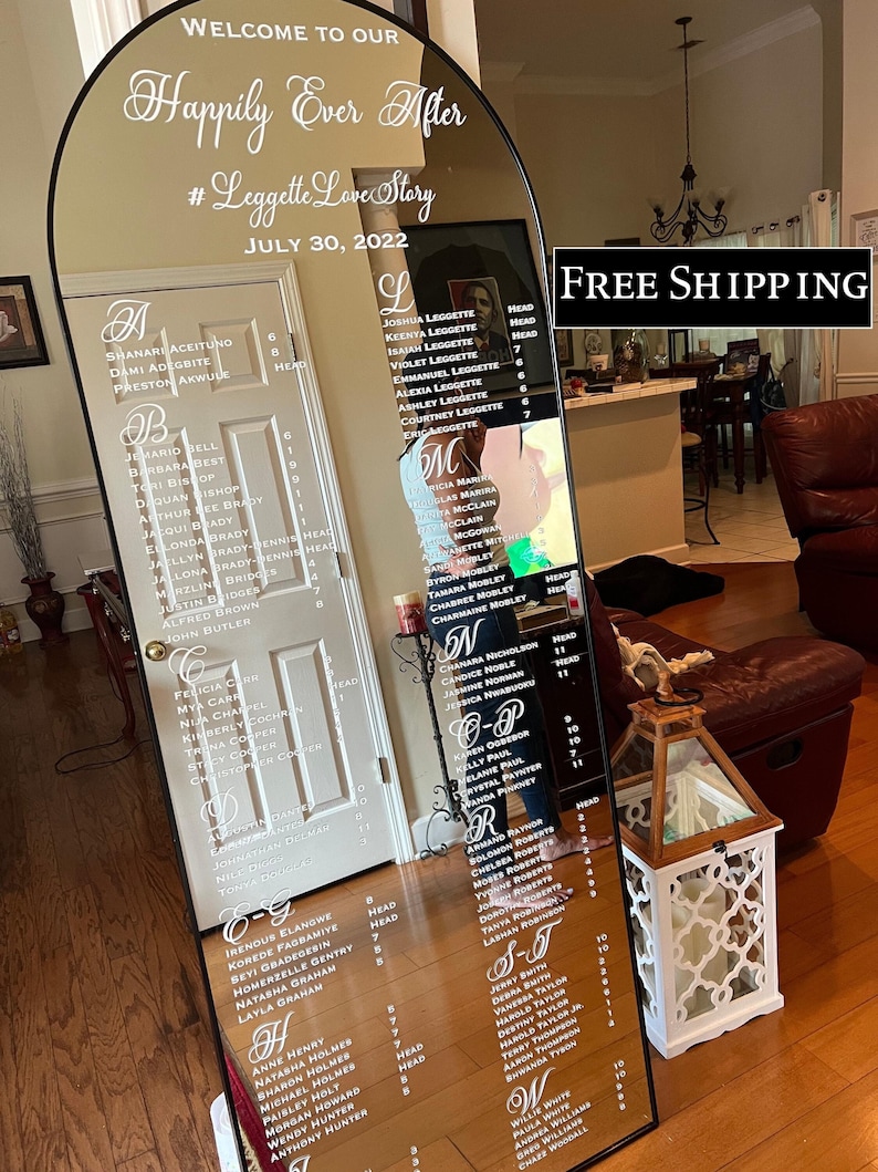 May include: An arched floor mirror featuring white text that reads Welcome to our Happily Ever After, #LeggetteLoveStory, and July 30, 2022. The mirror displays an alphabetical guest seating chart with names and table numbers printed in white.