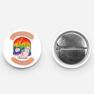 May include: A white button with a rainbow skull design and the text "I've got Rainbows in my bones". The back of the button is silver.
