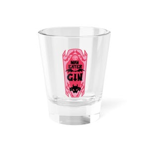 Puede incluir: Un vaso de chupito transparente con un diseño en forma de ataúd rosa y negro. El diseño presenta las palabras "MAN EATER GIN" en negro, con gráficos de murciélagos y demonios. El vaso está vacío y se encuentra sobre una superficie blanca.