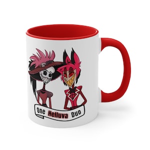 Puede incluir: Taza de cerámica blanca con interior y asa rojos. La taza presenta una ilustración de dibujos animados de dos personajes con el texto "One Helluva Duo" en una pancarta. Los personajes son en tonos rojos, negros y blancos.