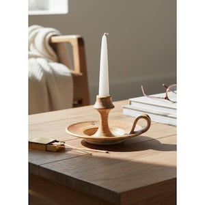 May include: A tan ceramic candle holder with a wide circular base, a central stem, and a curved side handle. A single white taper candle is inserted into the holder. The ceramic has a mottled, textured finish in shades of light and dark brown.