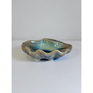 May include: A ceramic bowl shaped like a clam shell with a wavy, scalloped rim. The glaze features a blend of deep blue, turquoise, and earthy tan tones with a glossy finish. The bowl is supported by small, integrated feet on the underside.