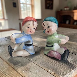 May include: Two ceramic figurines shaped like seated children with outstretched arms. One figure wears a red cap and blue tunic, while the other wears a teal cap and green tunic. Both have black shoes and painted facial features with open hands.