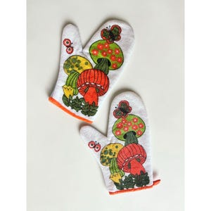 May include: A pair of white oven mitts featuring embroidered designs of orange and yellow mushrooms, green foliage, and small butterflies. The mitts have orange trim along the bottom edges and a textured fabric surface with colorful thread details.