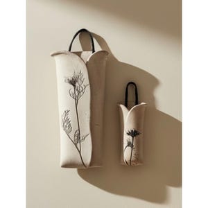 May include: Two off-white ceramic wall pockets with a matte finish, each featuring a black botanical line drawing of a wildflower. The vessels have a curved, petal-like opening and are suspended by a black cord loop attached to the top edge.