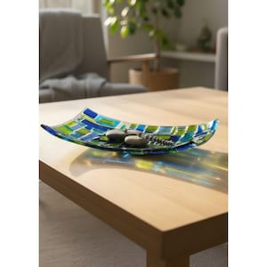 May include: A rectangular glass dish with a mosaic pattern of blue and green squares. The dish holds two smooth, gray stones and a string of small, green beads. The dish sits on a light-colored wooden table.