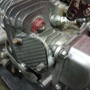 May include: Close-up of a motorcycle engine with a carbon fiber cover and a red spark plug cap. The engine is silver and has a black and white checkered pattern on the cover. The spark plug is labeled "SK".