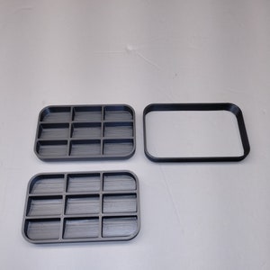 May include: Two gray plastic trays with compartments, one with a lid. The trays are designed to hold small items, such as jewelry or craft supplies.