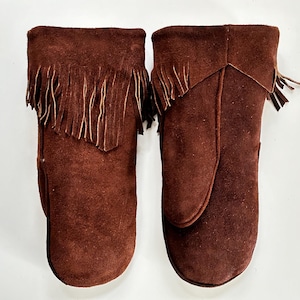May include: A pair of brown suede mittens with a fringed cuff. The mittens are a warm, earthy tone and appear to be made of soft suede material. The fringe adds a decorative element to the top of the mittens.