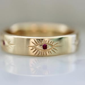 May include: A gold band ring with three ruby eyes engraved on the band. The eyes are surrounded by a sunburst design.