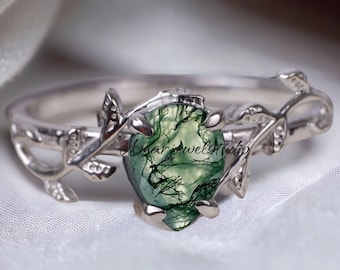 May include: A silver ring with a pear-shaped green moss agate stone. The ring features a delicate vine and leaf design on the band, with the stone set in a prong setting. The ring is displayed on a white, textured surface.