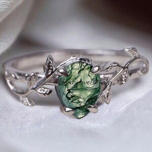 May include: A silver ring with a pear-shaped green moss agate stone. The ring features a delicate vine and leaf design on the band, with the stone set in a prong setting. The ring is displayed on a white, textured surface.