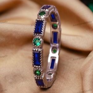 Könnte beinhalten: Ein silberner Bandring mit abwechselnd blauen und grünen rechteckigen Edelsteinen. Der Ring liegt auf einem goldenen Stoffhintergrund.