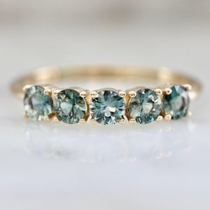 Puede incluir: Un anillo de banda de oro con cinco piedras preciosas redondas de color azul claro engastadas en una fila.