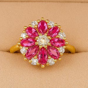 Puede incluir: Un anillo de oro con un diseño floral. El anillo tiene una piedra central rodeada de gemas rosas y blancas. El anillo está engastado en una banda de oro y se presenta en una caja de joyería.