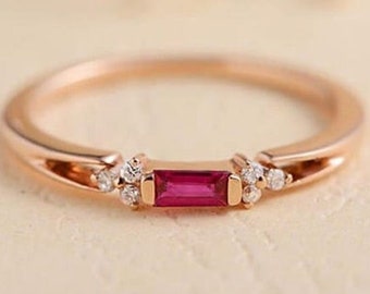 May include: A delicate rose gold ring with a small, rectangular ruby gemstone and smaller clear gemstones on either side.
