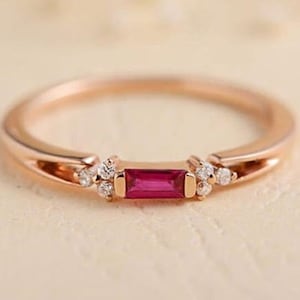 May include: A delicate rose gold ring with a small, rectangular ruby gemstone and smaller clear gemstones on either side.