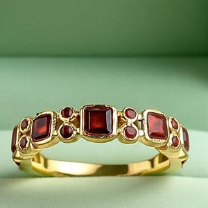 May include: A gold ring set with square and round red gemstones, presented in a light green velvet jewellery box. The background is a soft, blurred green.