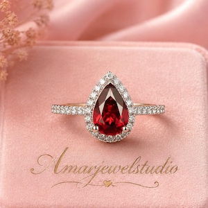 May include: A pear-shaped red gemstone ring featuring a halo of small white stones set on a thin metallic band. The ring rests on a square pink velvet box with the text Amarjewelstudio printed in gold script on the top surface.