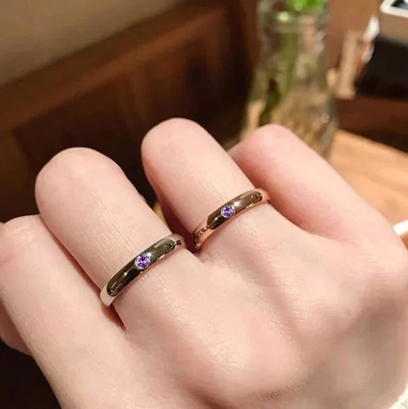 May include: Two simple silver and rose gold bands with a small purple gemstone on each.