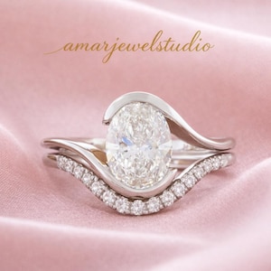 May include: A two-piece engagement ring set featuring a large oval-cut diamond and a curved band with smaller diamonds. The rings are set in a silver metal and rest on a pink satin surface. The text "amarjewelstudio" is in the upper left corner.
