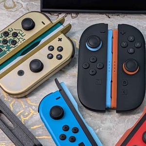 以下が含まれることがあります： 緑、赤、青、黒のJoy-Conを含む、カラフルなゲームコントローラーのコレクション。背景には黒いコンソールがあります。コントローラーはさまざまなデザインとボタン配置があり、ゲームをテーマにしています。