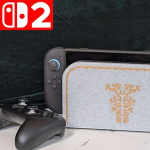 Könnte beinhalten: Eine schwarze Nintendo Switch-Konsole mit einer grau-gold gemusterten Hülle, einem schwarzen Controller und einer Figur. Die Hülle hat ein goldenes Design. Das Nintendo Switch-Logo befindet sich oben links.