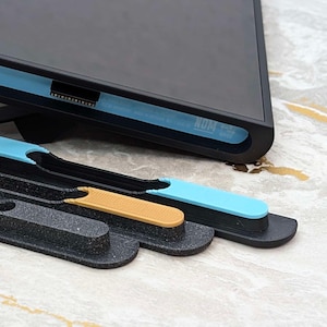 May include: A black electronic device with a blue accent, resting on a black stand. Three black trays hold gold and blue tools. The items are on a light-colored, patterned surface.
