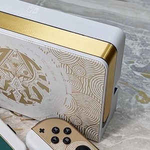 Può includere: Stazione di aggancio e controller Joy-Con Nintendo Switch bianchi e dorati. La stazione di aggancio presenta dettagli dorati e un design oro e bianco con motivi intricati. I controller Joy-Con sono oro e bianchi con pulsanti neri e accenti verdi.
