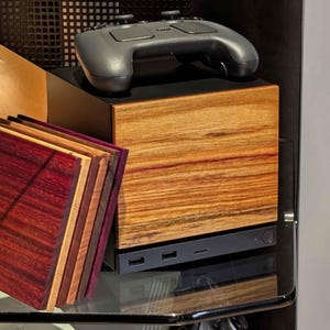 May include: A square wooden box with a light brown wood grain front panel and a black top, featuring a black game controller resting on top. Beside it stands a stack of four square wooden coasters in shades of deep purple, red, and brown.