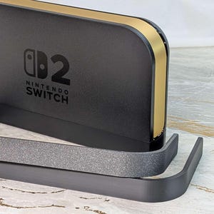 以下が含まれることがあります： 黒色のNintendo Switchドック。金色のトリムとNintendo Switchのロゴが付いています。ドックの前には、別の灰色のスタンドがあります。ゲーマー向けの洗練されたモダンなデザインです。