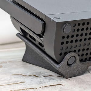 May include: A black Nintendo Switch console resting on a dark gray 3D-printed stand. The console is angled slightly upward, revealing the ventilation holes and a power button. The stand has a curved design with a circular support.