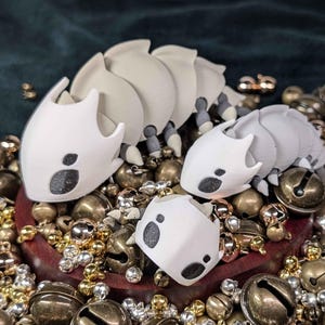 May include: A collection of white and gray segmented insect-like figurines with black markings, surrounded by a mix of gold, silver, and bronze bells. The figurines are arranged on a dark wooden base, set against a dark green background.