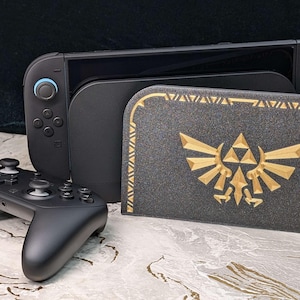 Könnte beinhalten: Eine schwarze Nintendo Switch-Konsole, ein schwarzer Controller und eine schwarze Hülle mit goldenem Emblem. Die Hülle zeigt ein goldenes Emblem mit Dreieck und Flügeln. Konsole und Controller liegen auf einer marmorähnlichen Oberfläche.