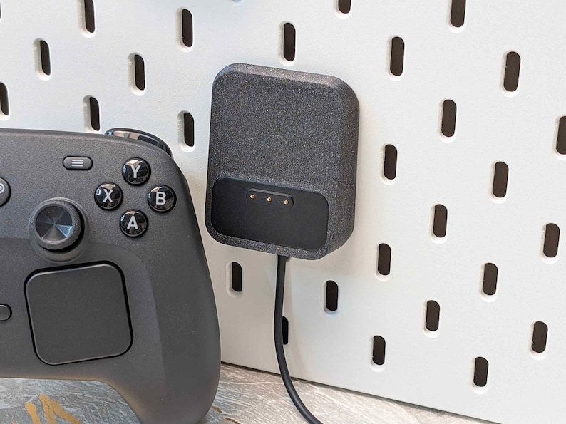 May include: A black video game controller next to a dark gray charging dock. The charging dock is mounted on a white pegboard. The controller has buttons labeled A, B, X, and Y.