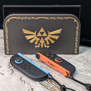 A photograph of a Zelda style dock cover / faceplate for the Nintendo Switch 2, based on Hylian Crest designs from the Legend of Zelda series. The dock cover has a black glitter theme with silky gold colored highlights in the shape of the Hyrule crest. The dock cover is fitted on the Nintendo Switch 2 dock, and the Switch 2 Joycons are in front of the dock.