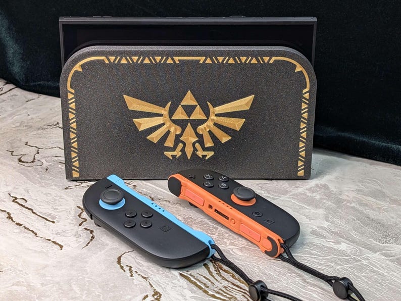 A photograph of a Zelda style dock cover / faceplate for the Nintendo Switch 2, based on Hylian Crest designs from the Legend of Zelda series. The dock cover has a black glitter theme with silky gold colored highlights in the shape of the Hyrule crest. The dock cover is fitted on the Nintendo Switch 2 dock, and the Switch 2 Joycons are in front of the dock.