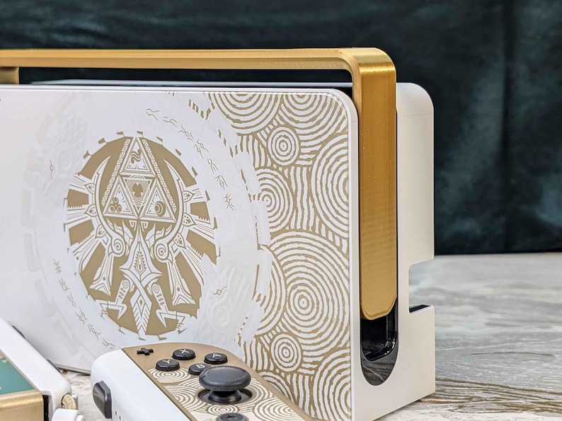 May include: White and gold video game console with matching controllers. The console features a gold trim and a white surface with a gold circular pattern and a central emblem. The controllers have gold accents and black buttons.