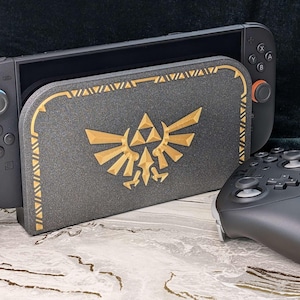 A photograph of a Zelda style dock cover / faceplate for the Nintendo Switch 2, based on designs from the Legend of Zelda series. The dock cover has a black glitter theme with silky gold colored highlights in the shape of the Hyrule crest. The dock cover is fitted on the Nintendo Switch 2 dock, and the Switch 2 Pro Controller is in front of the dock.