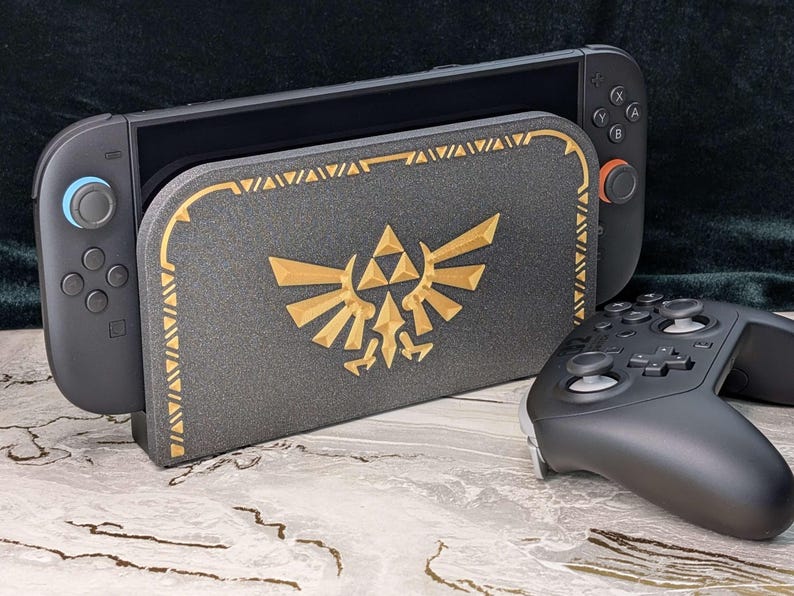 A photograph of a Zelda style dock cover / faceplate for the Nintendo Switch 2, based on designs from the Legend of Zelda series. The dock cover has a black glitter theme with silky gold colored highlights in the shape of the Hyrule crest. The dock cover is fitted on the Nintendo Switch 2 dock, and the Switch 2 Pro Controller is in front of the dock.