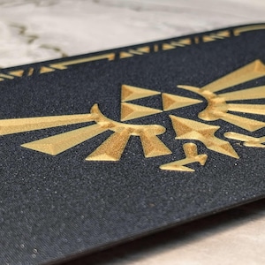 A closeup photograph of a Zelda style dock cover / faceplate for the Nintendo Switch 2, based on Crest of Hyrule designs from the Legend of Zelda series. This closeup photo shows a detailed view of the Hylian Crest in a galaxy black glitter finish with silky gold colored highlights.