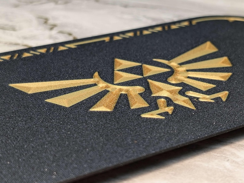 A closeup photograph of a Zelda style dock cover / faceplate for the Nintendo Switch 2, based on Crest of Hyrule designs from the Legend of Zelda series. This closeup photo shows a detailed view of the Hylian Crest in a galaxy black glitter finish with silky gold colored highlights.