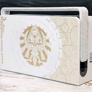 May include: White and gold decorative gaming console stand with a handle. The stand features a gold emblem and circular patterns on the front panel. The handle and side panels are white with a speckled texture. The design is inspired by a popular video game.