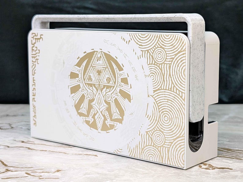 May include: White and gold decorative gaming console stand with a handle. The stand features a gold emblem and circular patterns on the front panel. The handle and side panels are white with a speckled texture. The design is inspired by a popular video game.