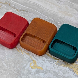 May include: Three rectangular, open-top containers in red, brown, and teal. The containers have a recessed area on one side. The brown container has a textured, honeycomb-like pattern. The containers are arranged side by side.