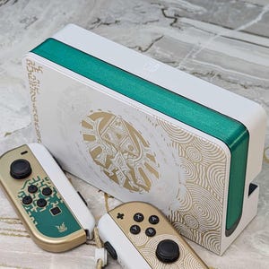 May include: White and teal gaming console with matching controllers. The console features gold accents and a teal stripe. The controllers are gold and white with teal and black buttons. The console and controllers have a decorative design.