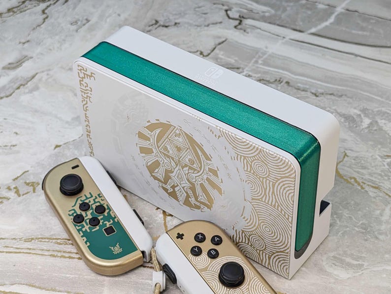 May include: White and teal gaming console with matching controllers. The console features gold accents and a teal stripe. The controllers are gold and white with teal and black buttons. The console and controllers have a decorative design.