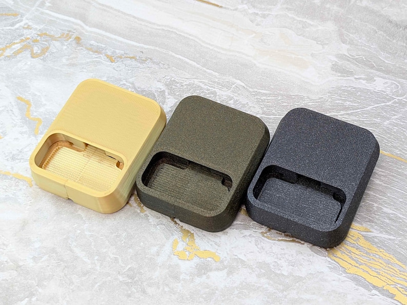 May include: Three rectangular, textured cases in a row. The cases are in shades of yellow, olive green, and dark gray. Each case has a rectangular cutout on one side. The cases are on a light-colored, patterned surface.